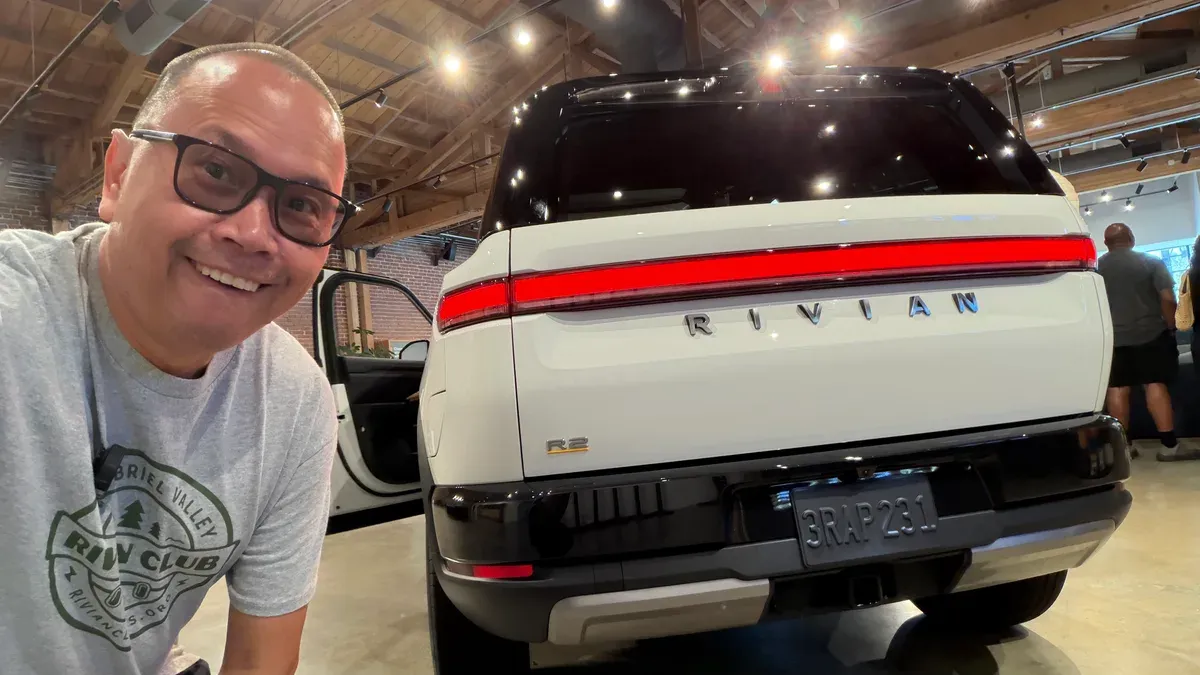 Sherwin with the rear of a white Rivian R2 showing the full-width taillight and R2 badge