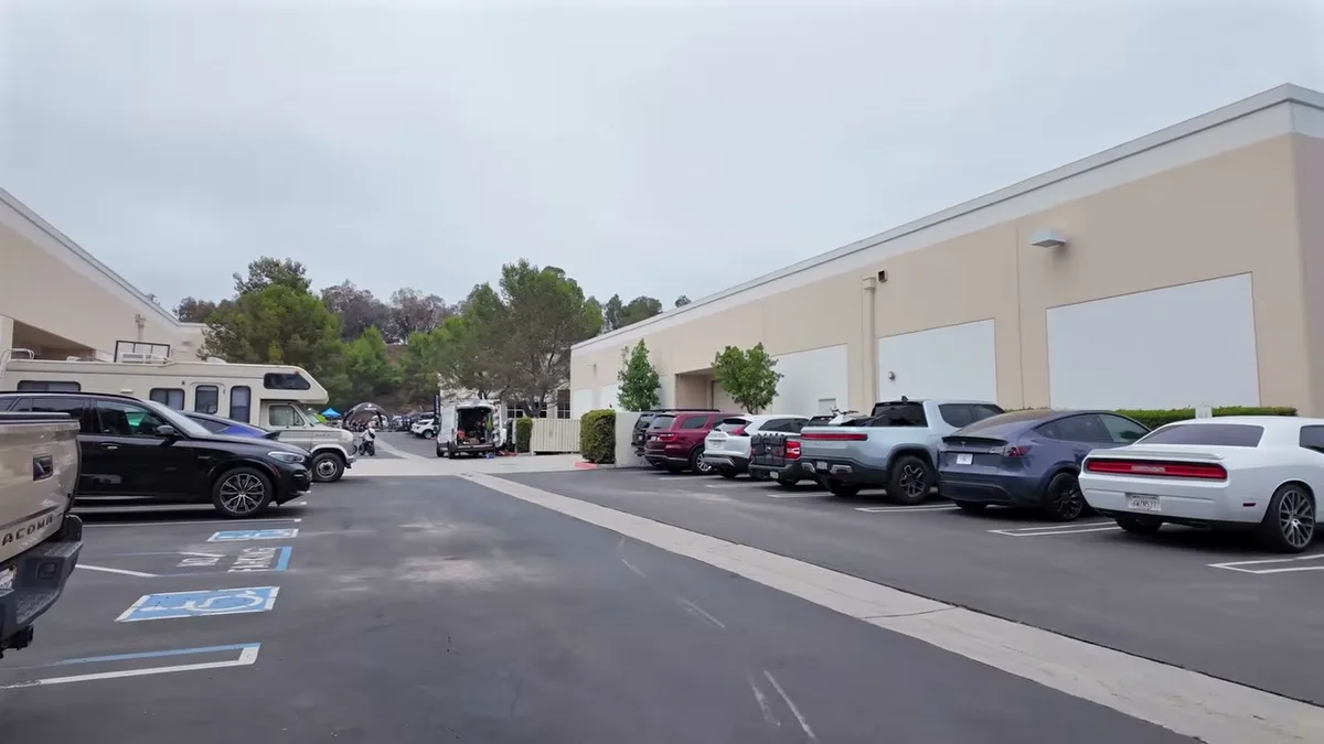 Wide shot of a parking lot with Rivian trucks, SUVs, a Dodge Challenger, and an RV outside a building.