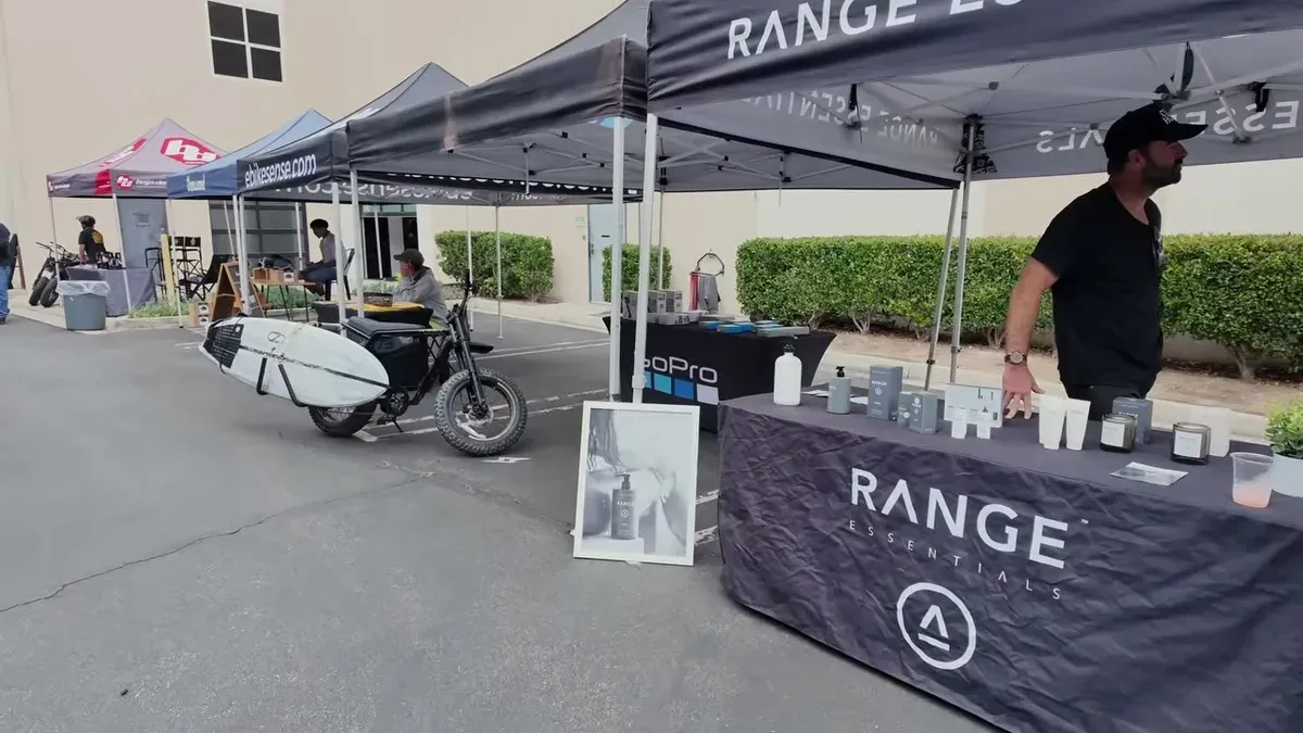 Outdoor event with "RANGE ESSENTIALS" booth, pop-up tents, and a Super73 electric bike with a surfboard.