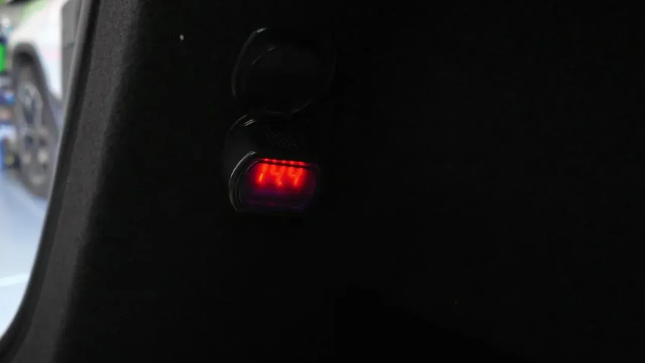 A cigarette-lighter voltmeter plugged into the 12V socket of a Tesla Model Y Juniper, its red display reading 14.4 volts.