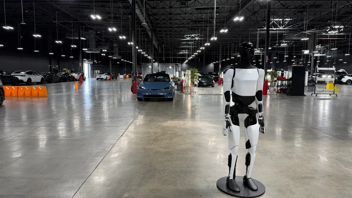Tesla Optimus humanoid robot standing on a display stand in the Montebello service center, with Teslas parked in the background.
