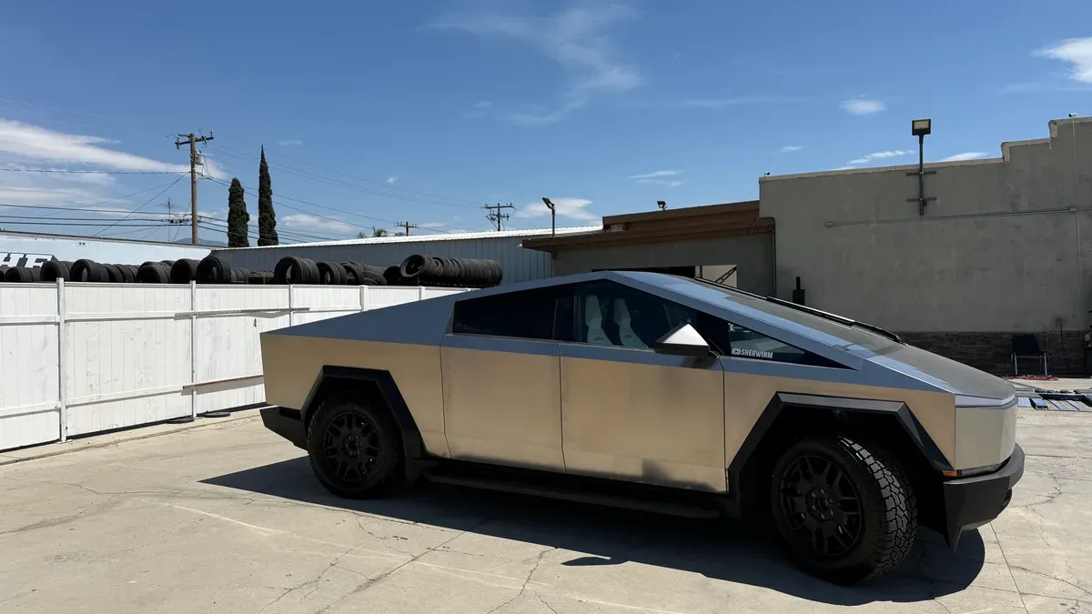 Tesla Cybertruck parked outdoors on a concrete lot next to a white fence under a blue sky.