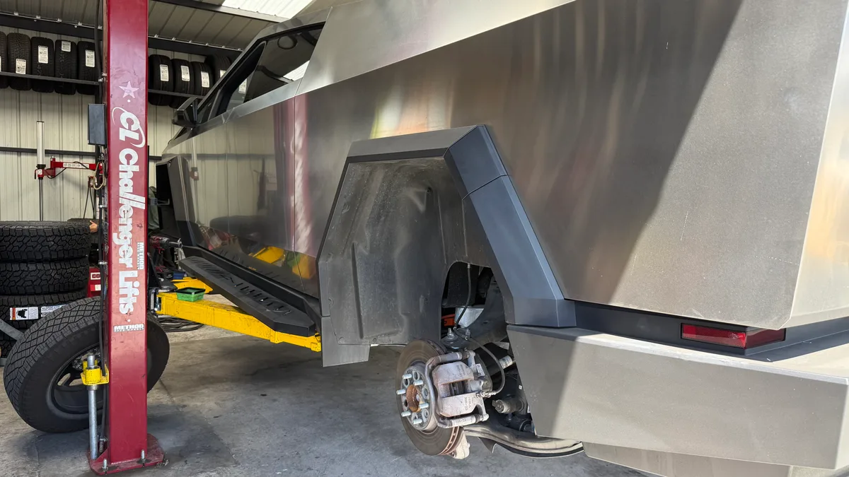 Tesla Cybertruck lifted on a yellow jack stand with the rear wheel removed, showing brake rotor and suspension.