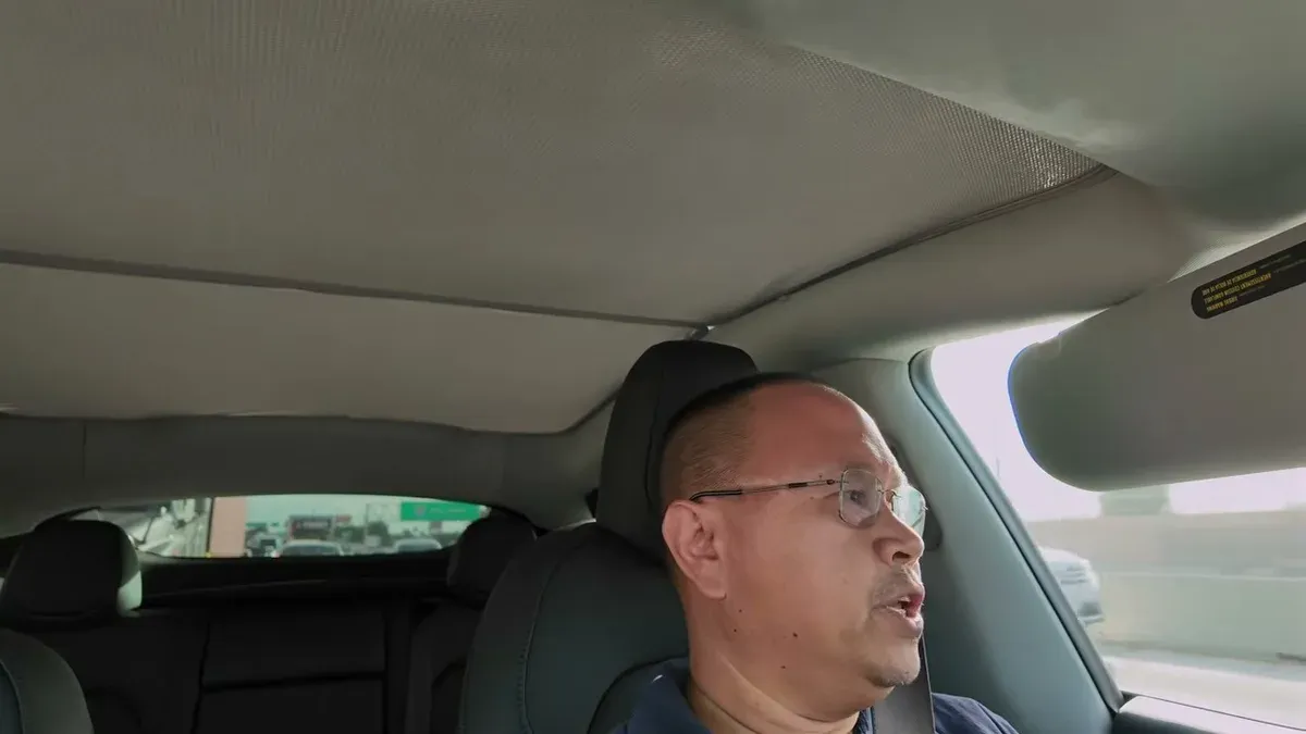 Man driving Tesla on highway with roof shade installed above him, residential buildings visible through windows