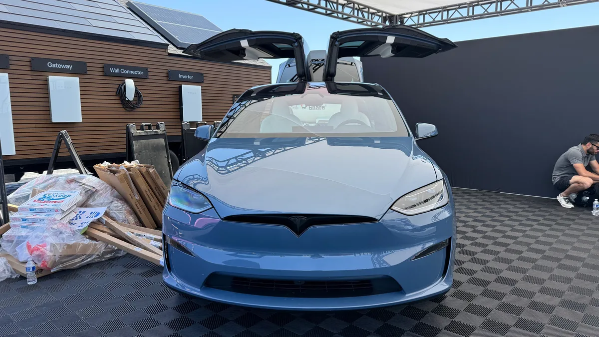 Light blue Tesla Model X with falcon-wing doors open in front of a Tesla home energy display featuring solar panels and a Wall Connector at Electrify Expo