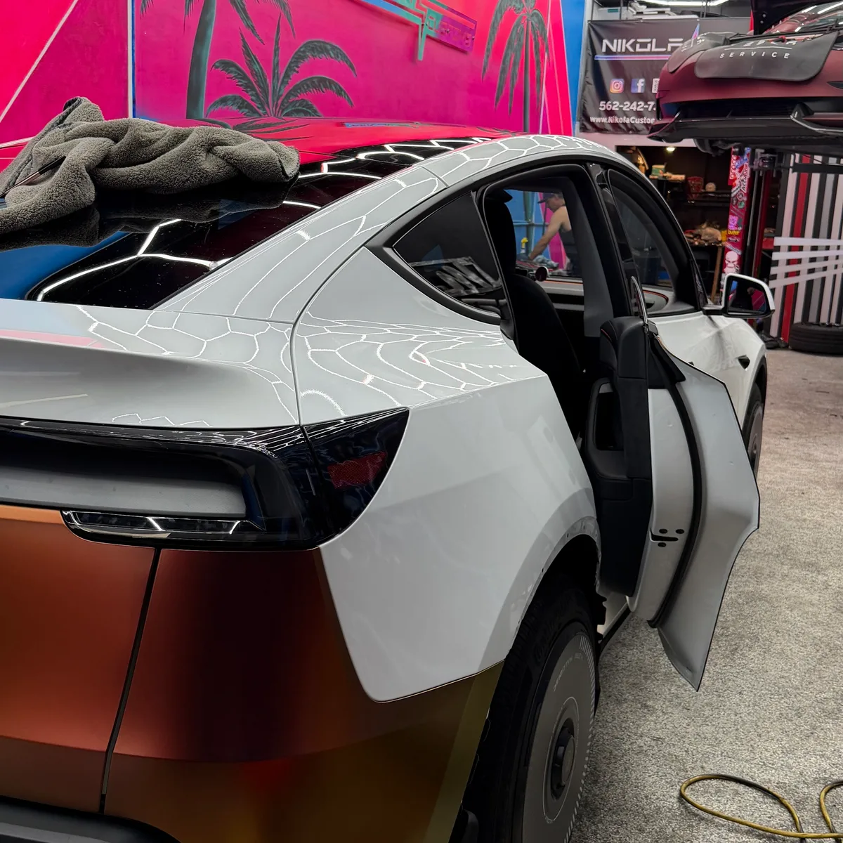 Tesla Model Y rear door open mid-wrap, bronze color visible on the quarter panel, pink palm tree mural in background