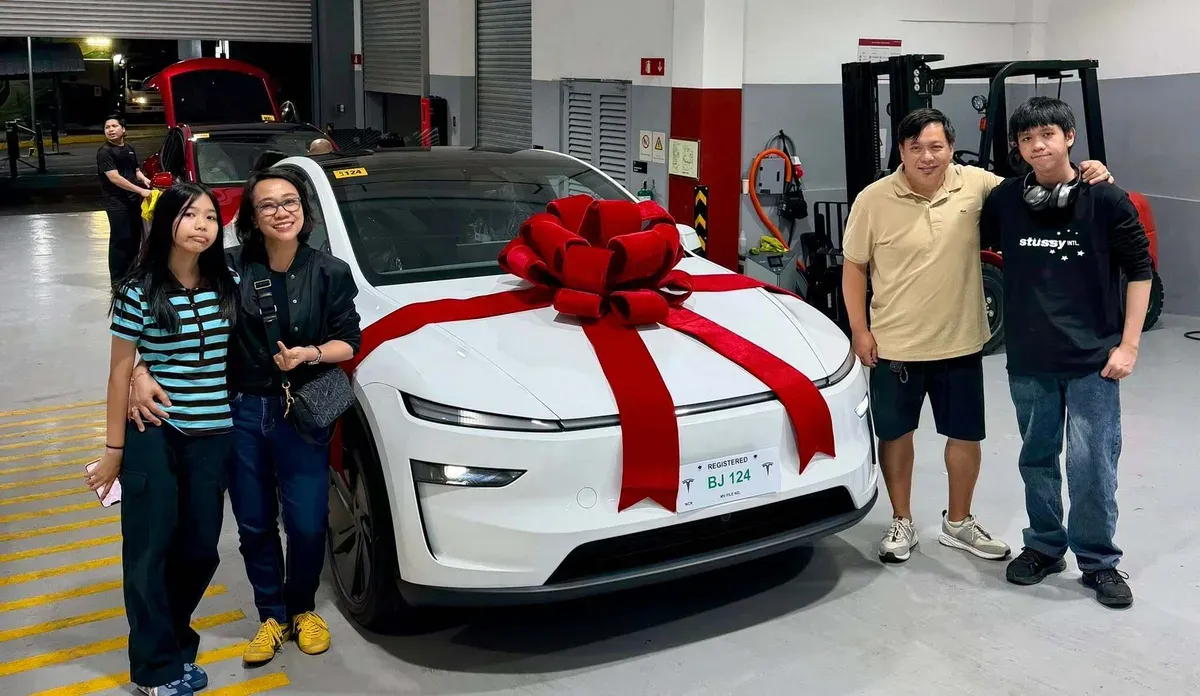 Joy and her family beside their new white Tesla Model Y with a large red bow on the hood at the Tesla delivery center in the Philippines