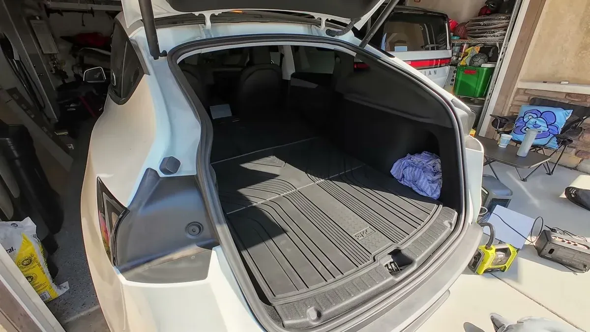 Open trunk of white Tesla Model Y with black liner and blue patterned pillow, ready for the air mattress