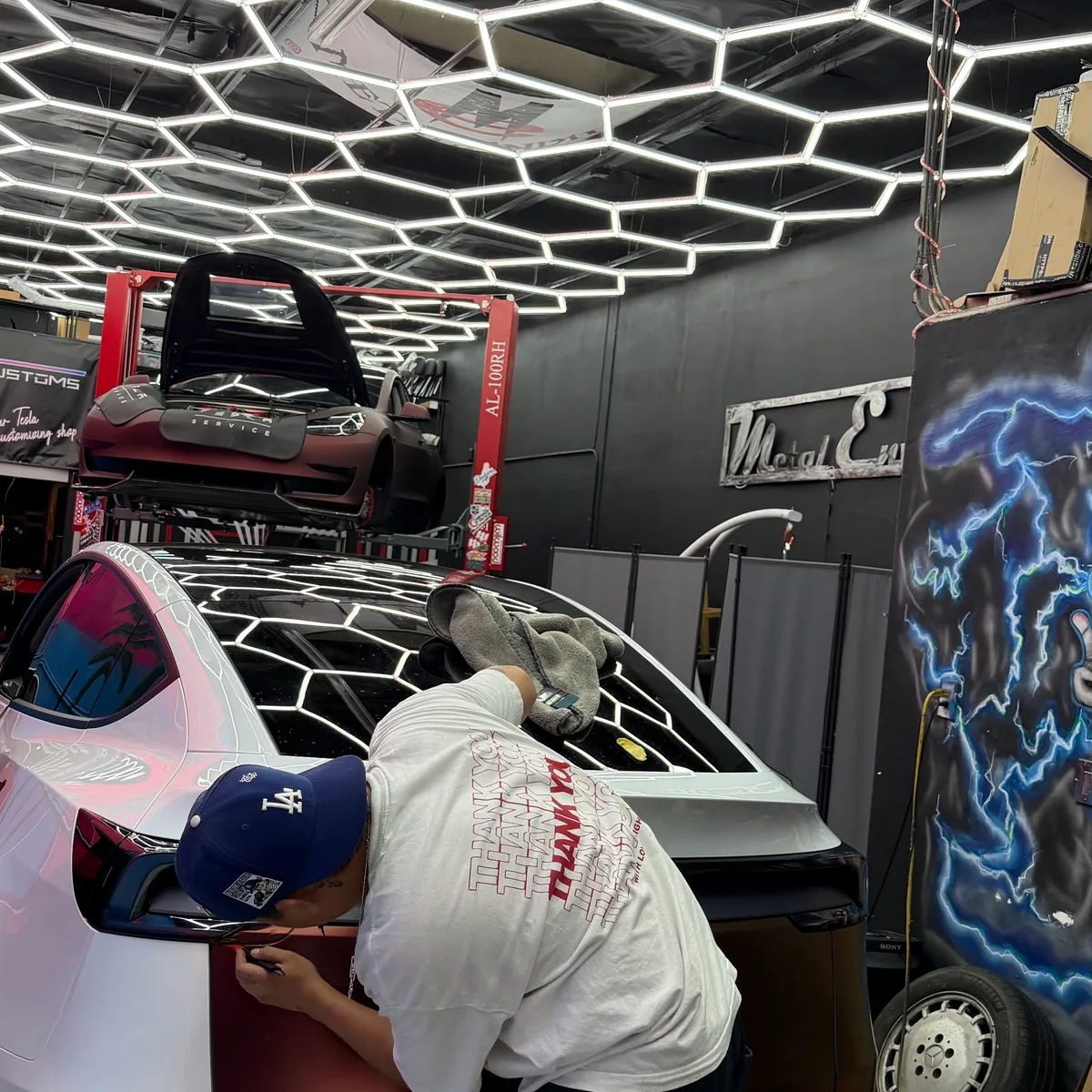 Technician applying Rising Sun vinyl wrap to a white Tesla Model Y inside Nikola Customs, hexagonal ceiling lights overhead