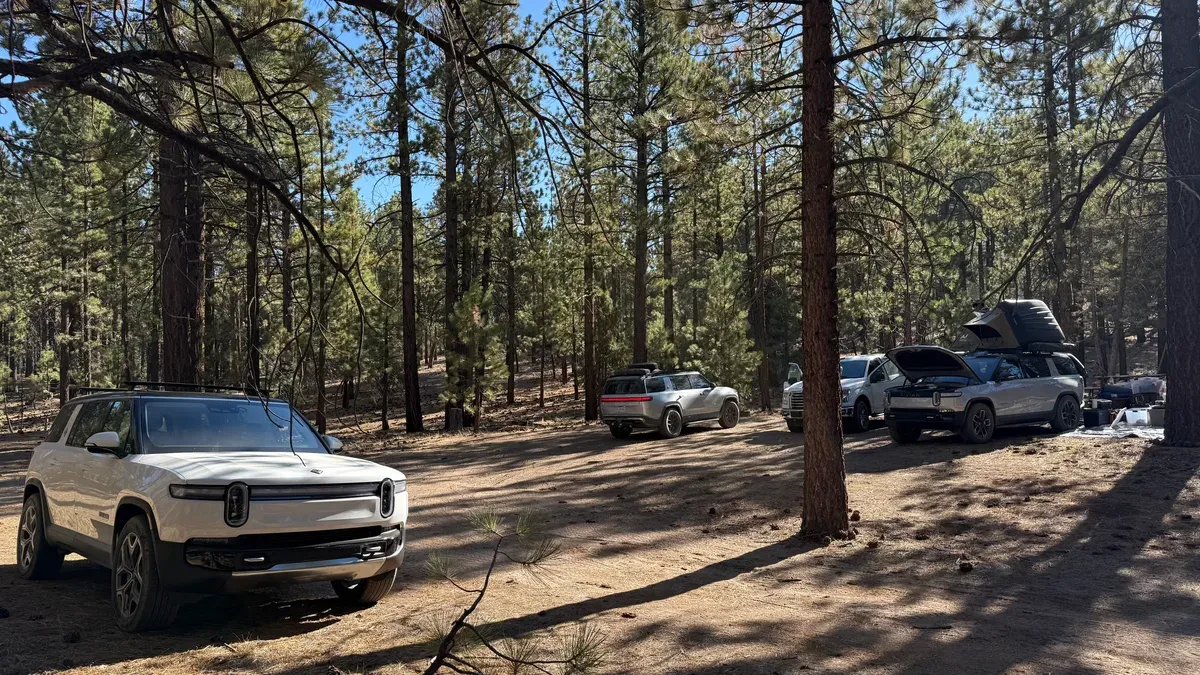 White Rivian R1S parked in foreground at Holcomb Valley pine forest campsite with other Rivians and camping setups