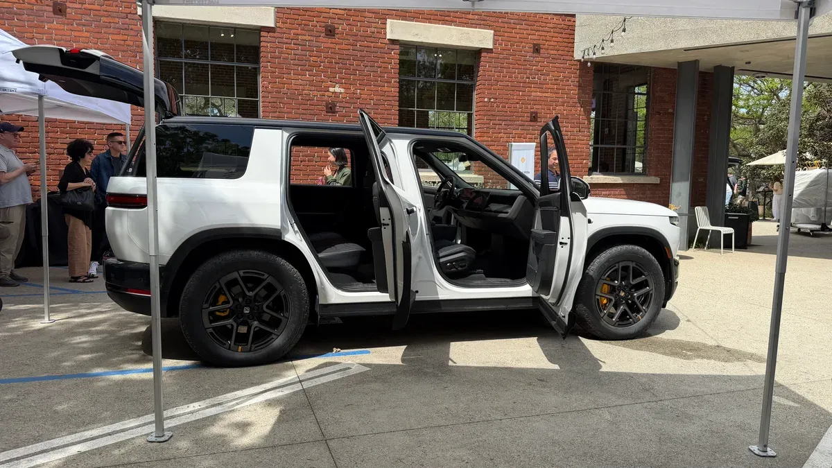 White Rivian R2 with all doors and trunk open at the outdoor event, showing black interior.