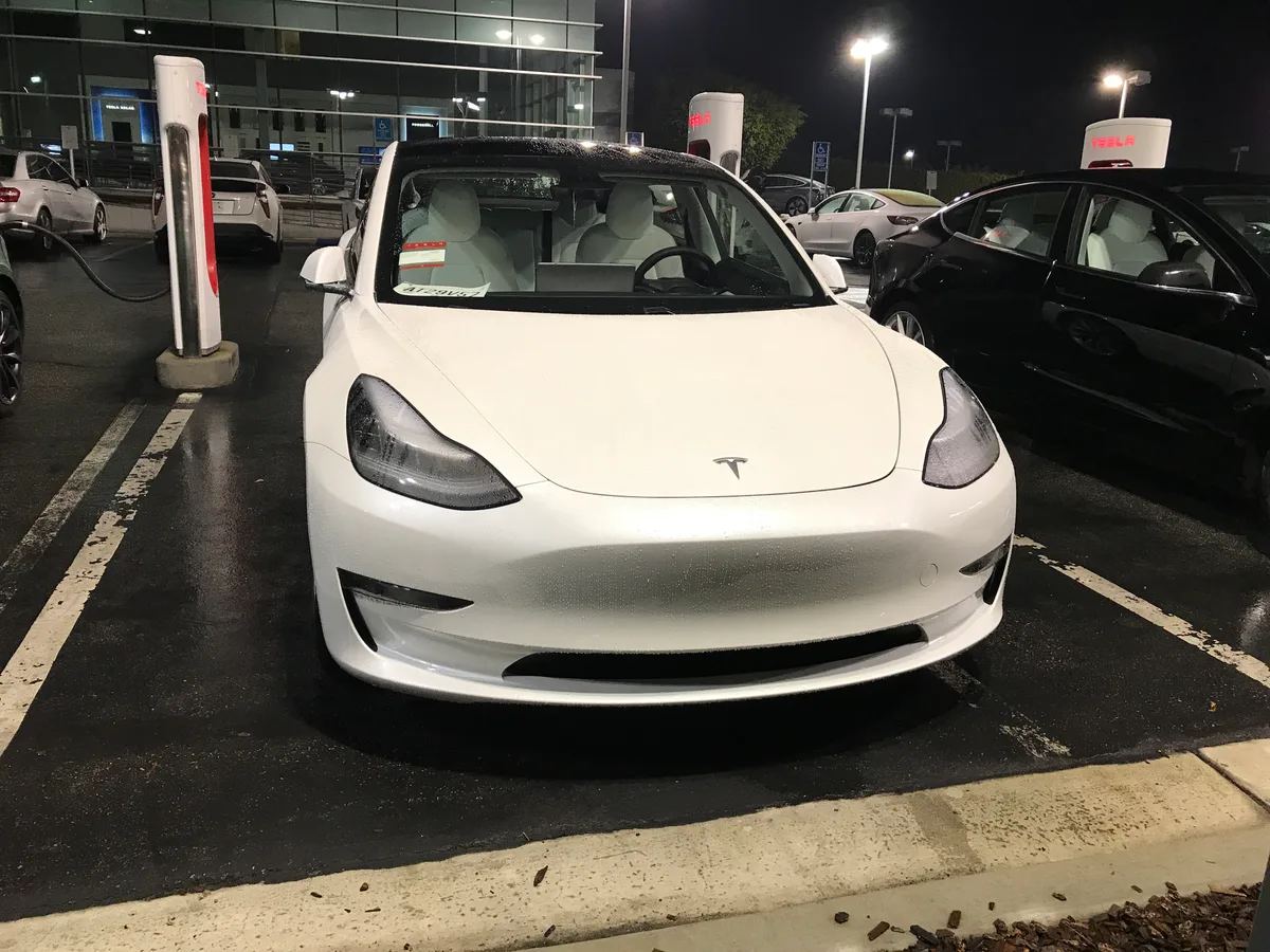 White 2020 Tesla Model 3 parked at the Buena Park Tesla dealership at night, rain on the ground