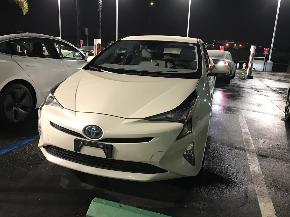 White 2016 Toyota Prius parked at the Tesla dealership lot at night, surrounded by Superchargers — the trade-in